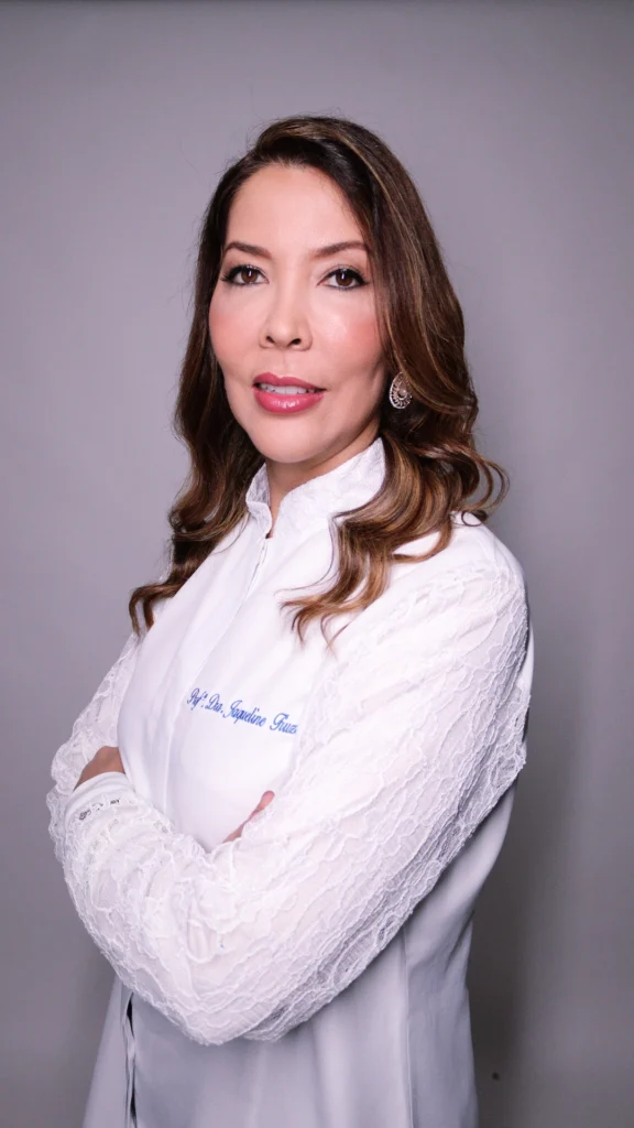 Dra. Jaqueline Fiuza, dentista em Valinhos, posa profissionalmente com os braços cruzados vestindo jaleco branco com detalhes em renda e seu nome bordado em azul, em um estúdio com fundo cinza