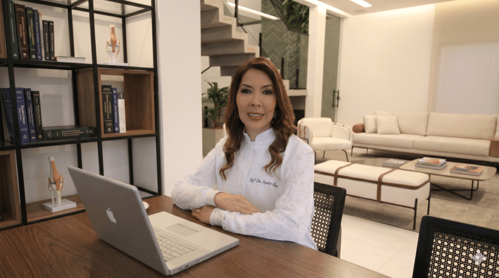 Drª Jaqueline Souza, especialista em preenchimento facial Valinhos, sorrindo profissionalmente em seu consultório moderno, com estante de livros e área de estar sofisticada ao fundo.